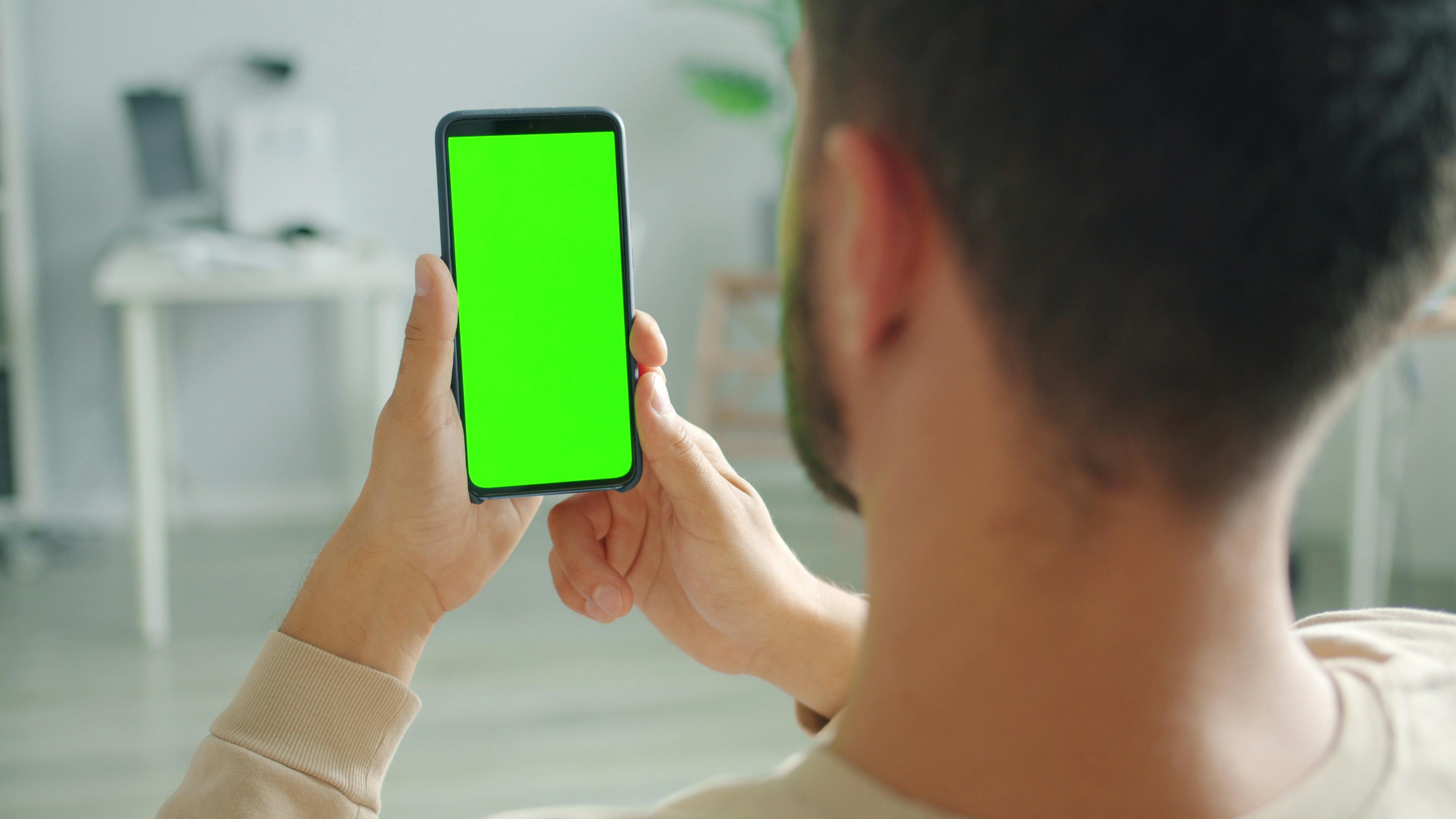 A person using a smartphone with a green screen in a home setting.