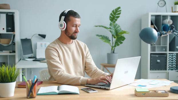 Adult man with headphones working on laptop in home office setting.
