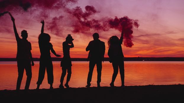 Five people joyfully celebrate at sunset by a lake with colorful smoke bombs.