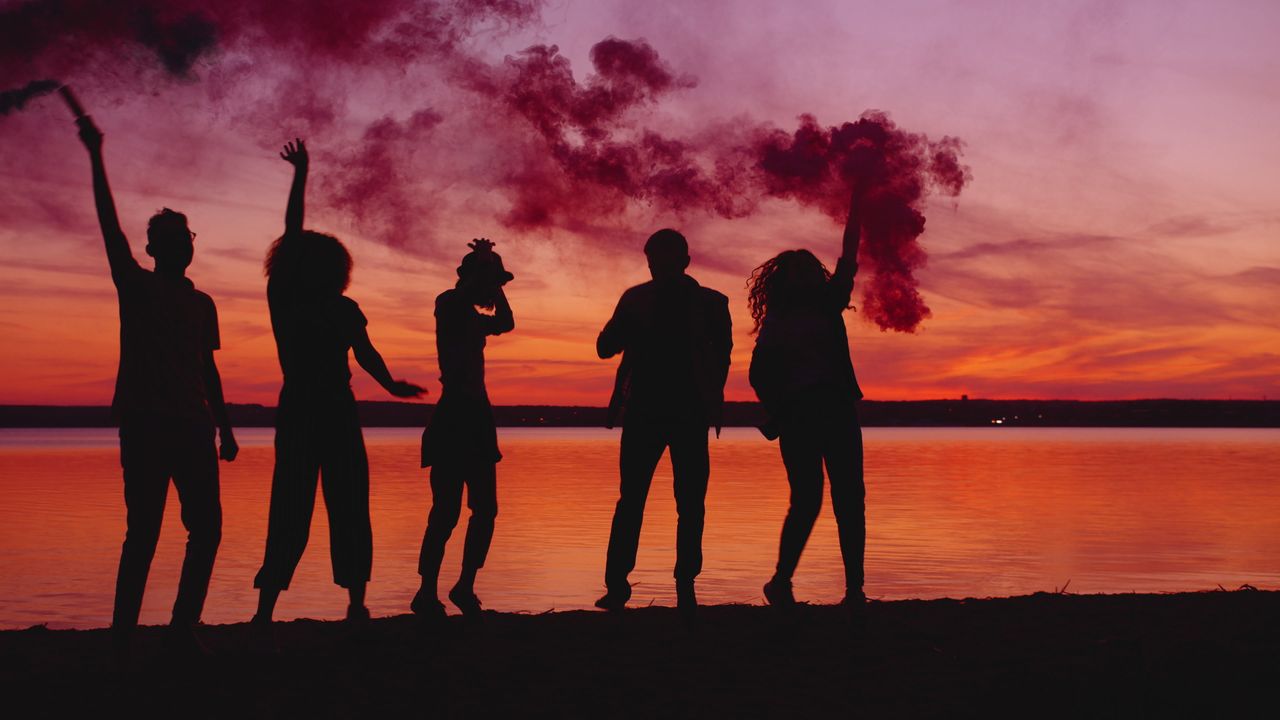 five people joyfully celebrate at sunset by a lake with colorful smoke
