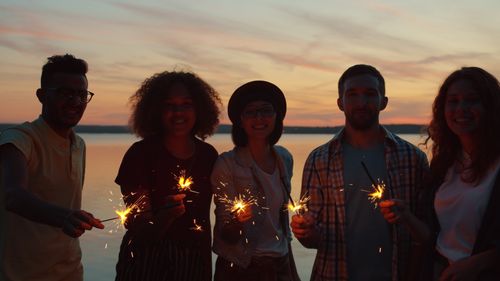 Free Friends holding sparklers by a lake during sunset, enjoying a festive evening. Stock Photo