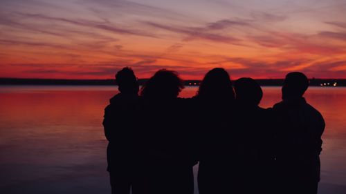 Free Silhouetted group of friends watching a colorful sunset over a serene lake. Stock Photo