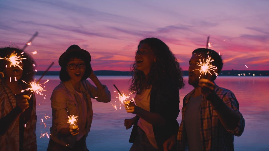 Group of friends having fun with sparklers by a lake at sunset.