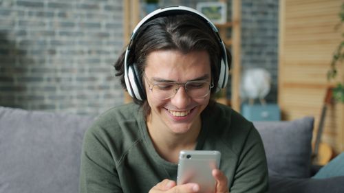 Free Young man with headphones smiling while using smartphone indoors. Stock Photo