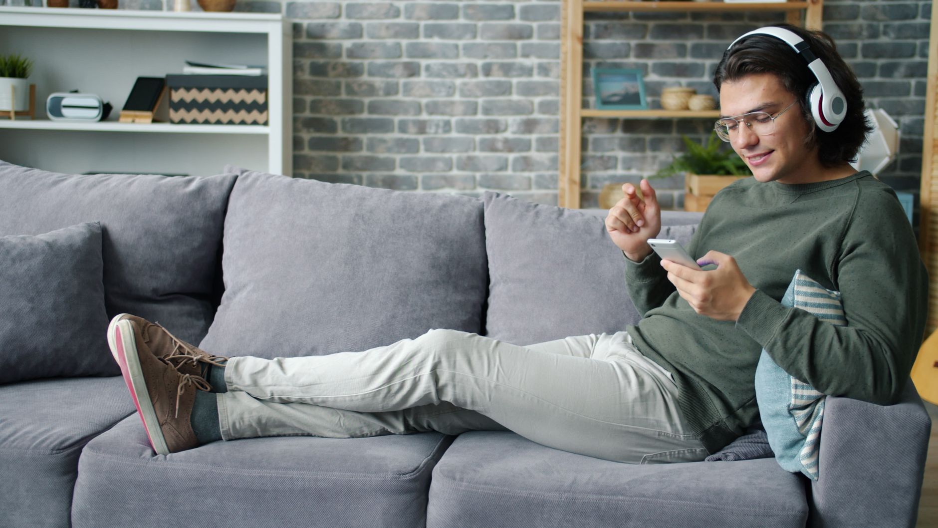 Casually dressed man listening to music on headphones while using a smartphone on a cozy couch.