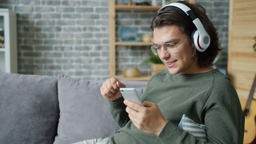 Free Casual young man wearing headphones, using smartphone, relaxing on a couch. Stock Photo