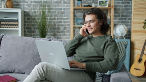 Man relaxing and working from home on a laptop while talking on a phone.