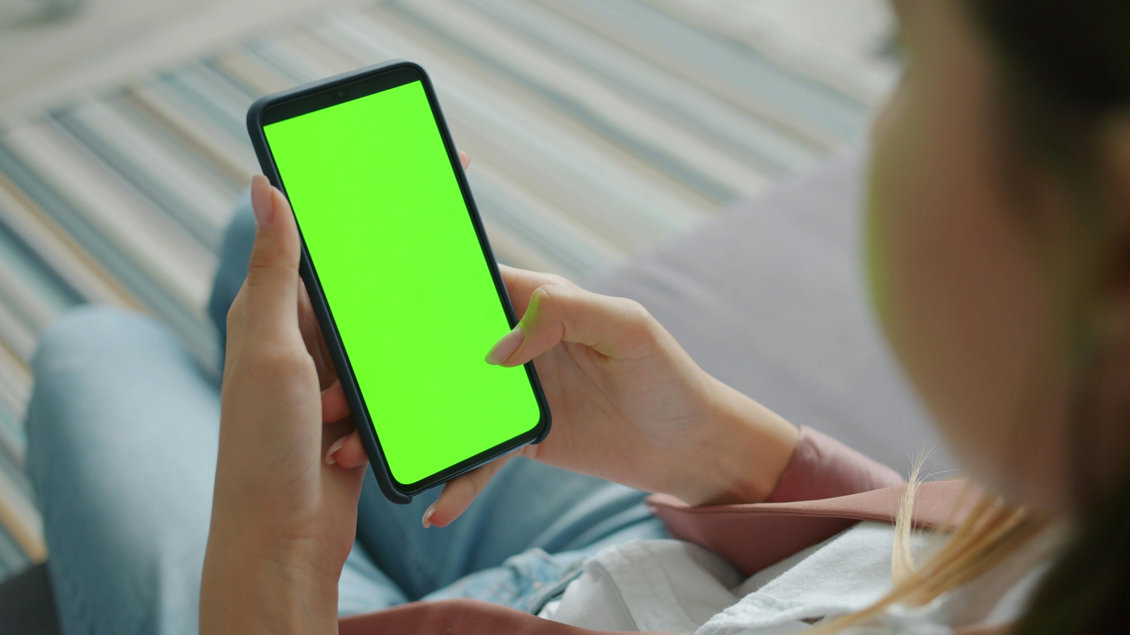 Close-up of person holding smartphone with green screen indoors, for digital overlay.