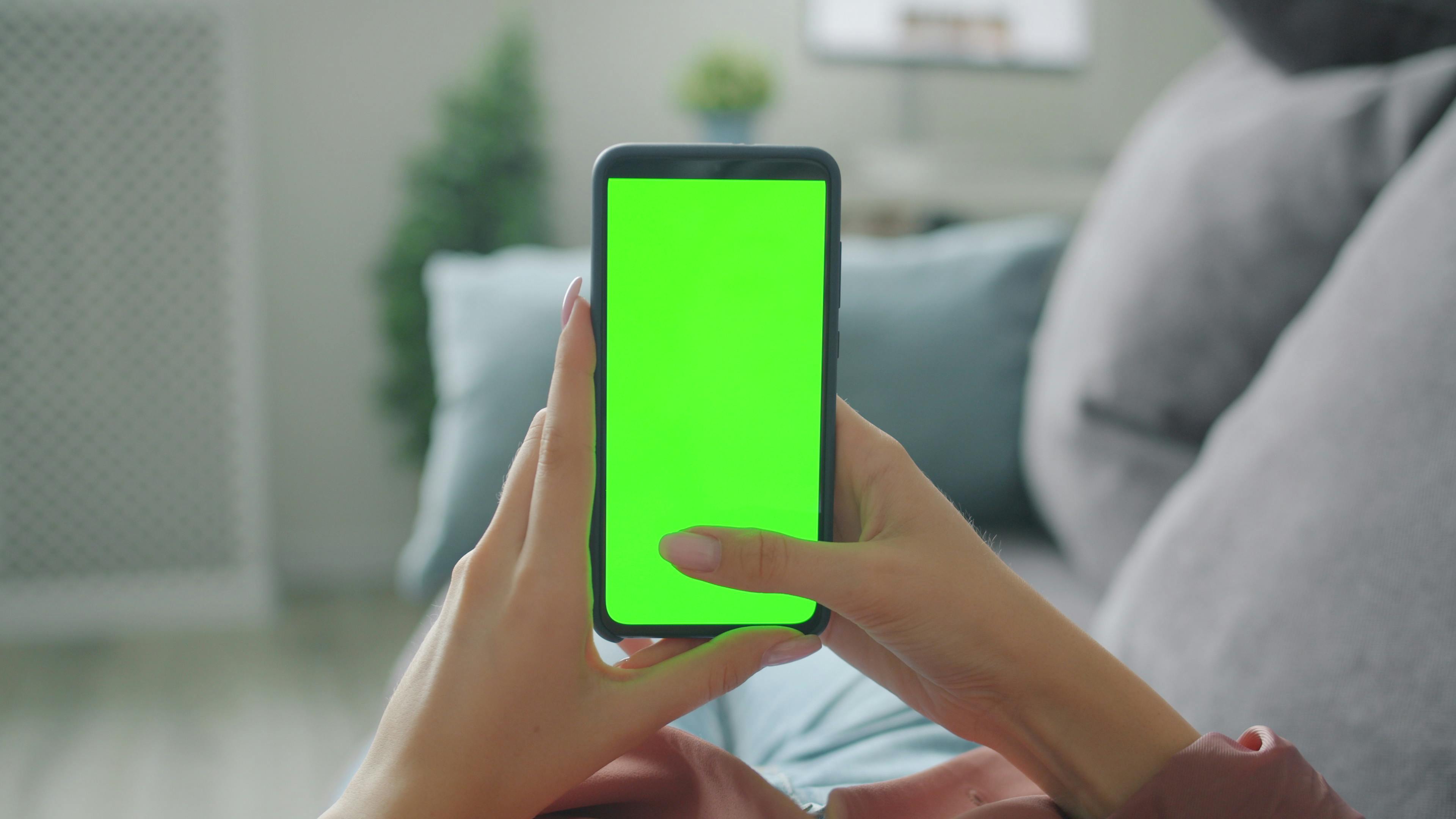 Person holding a smartphone with a blank green screen indoors on a sofa.