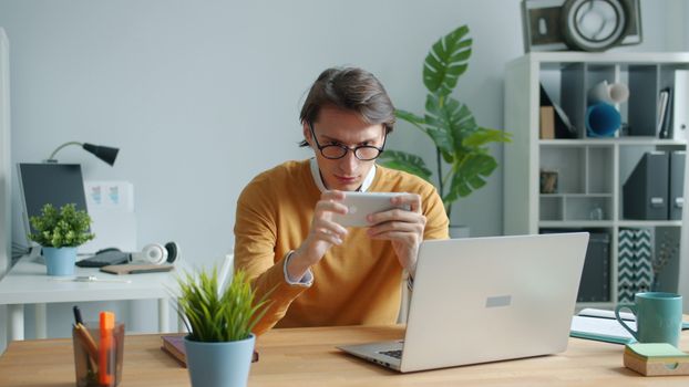 Man concentrating on mobile phone at work desk with laptop and plants, indoor setting.
