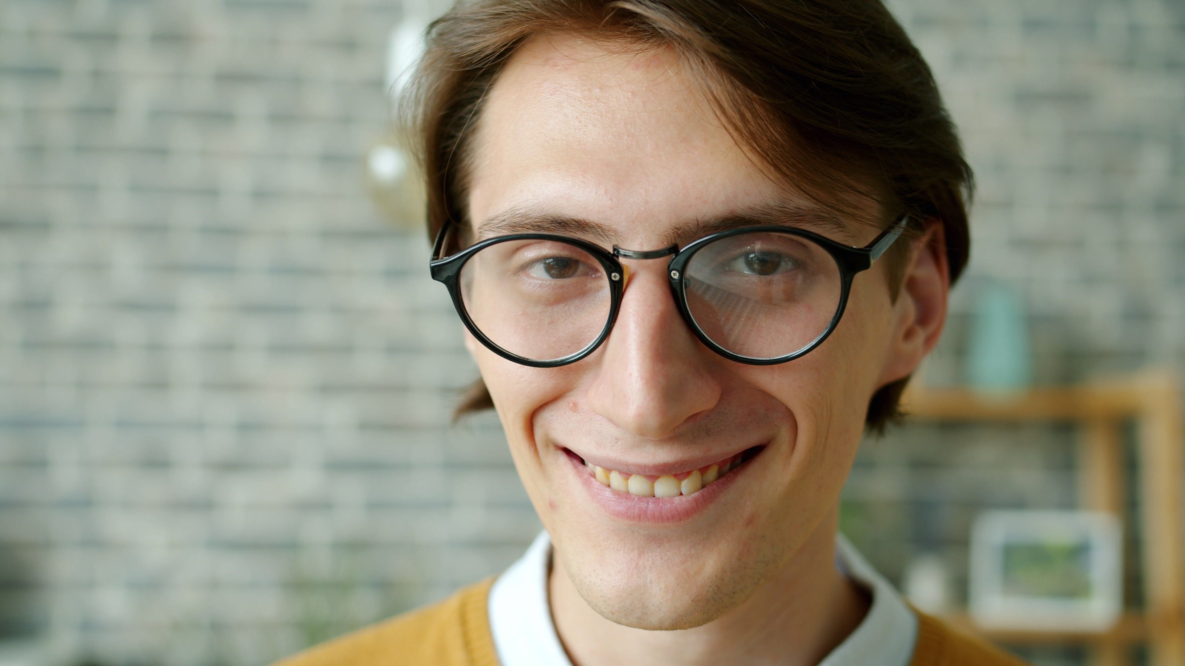 Close-up portrait of a smiling young man with glasses against brick wall indoors.
