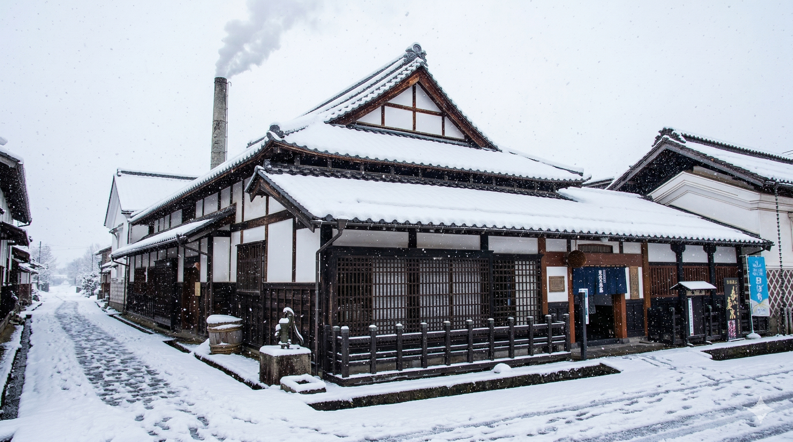 Snow-covered traditional Japanese sake brewery with chimney during winter snowfall.
