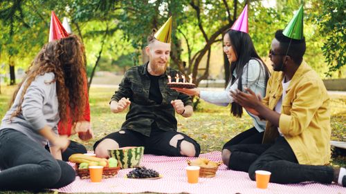 Free Friends celebrate a birthday outdoors with cake, hats, and picnic setup. Stock Photo