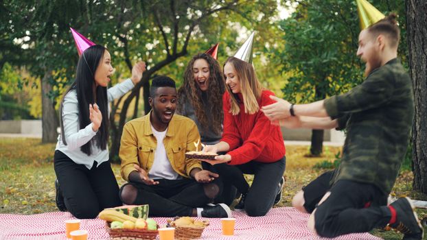 Friends celebrate a birthday with a picnic and cake outdoors, wearing party hats.