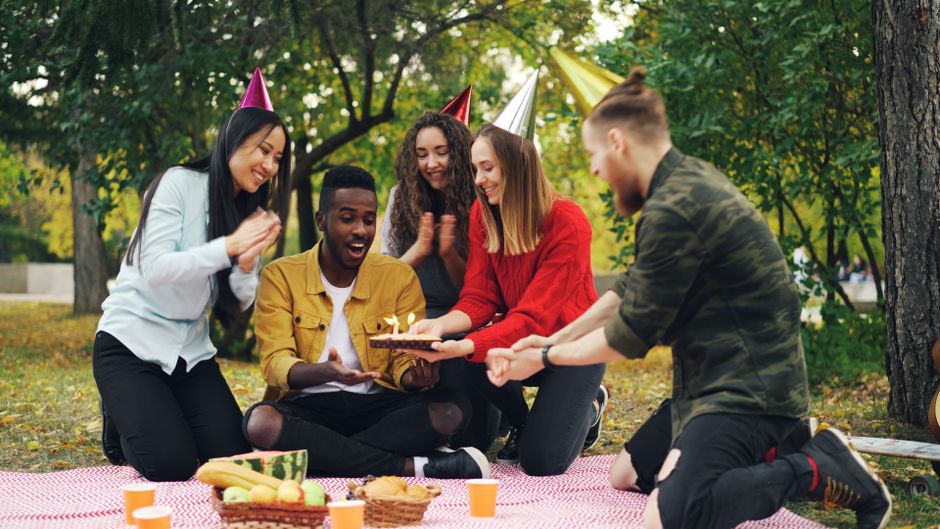 Group of friends enjoying a birthday celebration outdoors with cake and party hats.