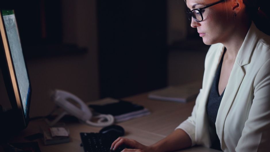 Mujer con gafas trabajando tarde en un escritorio con un monitor de computadora.