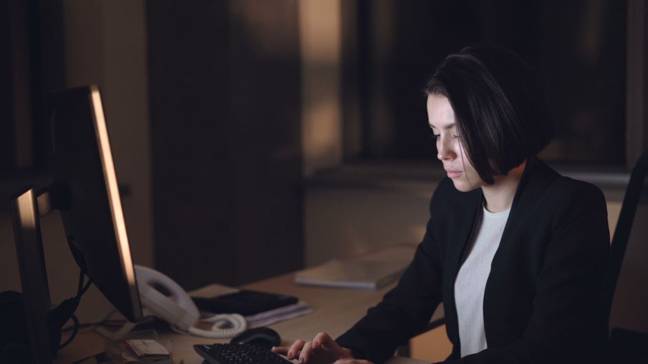 Una mujer vestida con traje trabaja tarde en un escritorio poco iluminado, concentrada en su computadora.