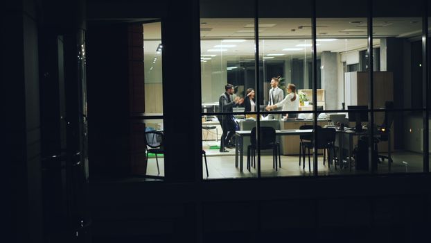 Business team celebrating success inside a modern glass office at night.