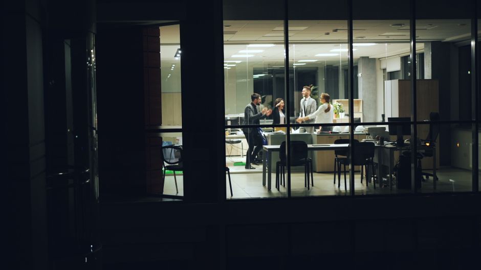 Business team celebrating success inside a modern glass office at night.