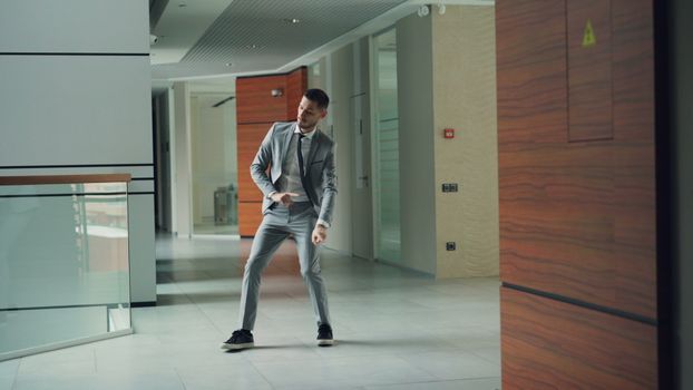 Young businessman in a suit dancing happily in a modern office corridor.