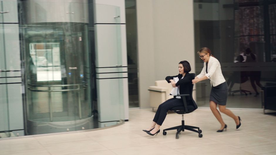 Dos empresarias se divierten jugando a las carreras de sillas de oficina en un vestíbulo moderno.
