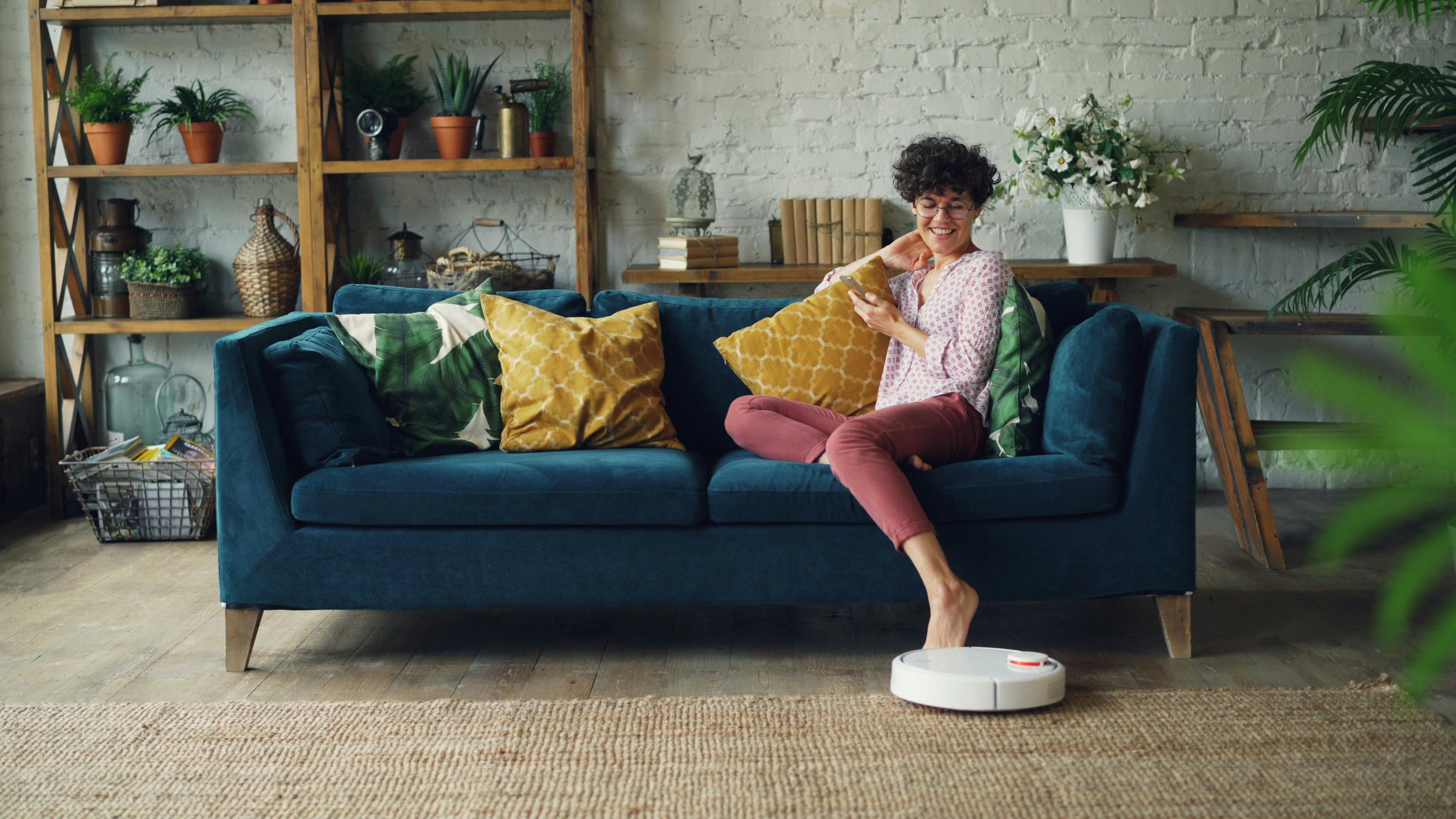 Woman relaxing on sofa while a robotic vacuum cleans the room, showcasing a cozy modern interior.