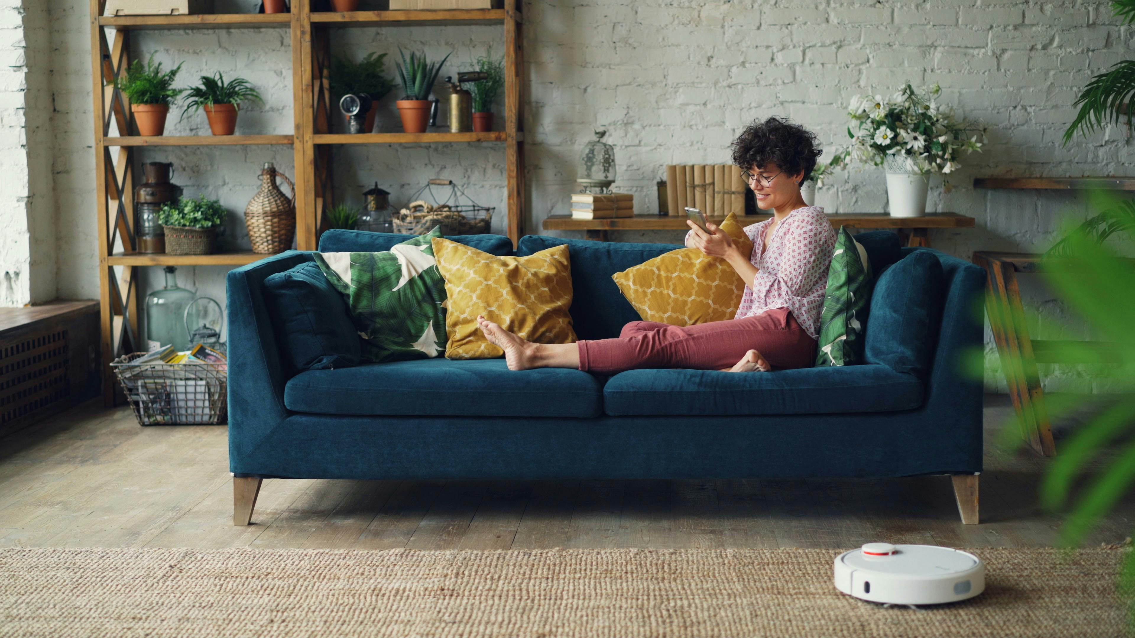 Adult enjoying a relaxing day on a cozy sofa with a phone and smart cleaning robot.