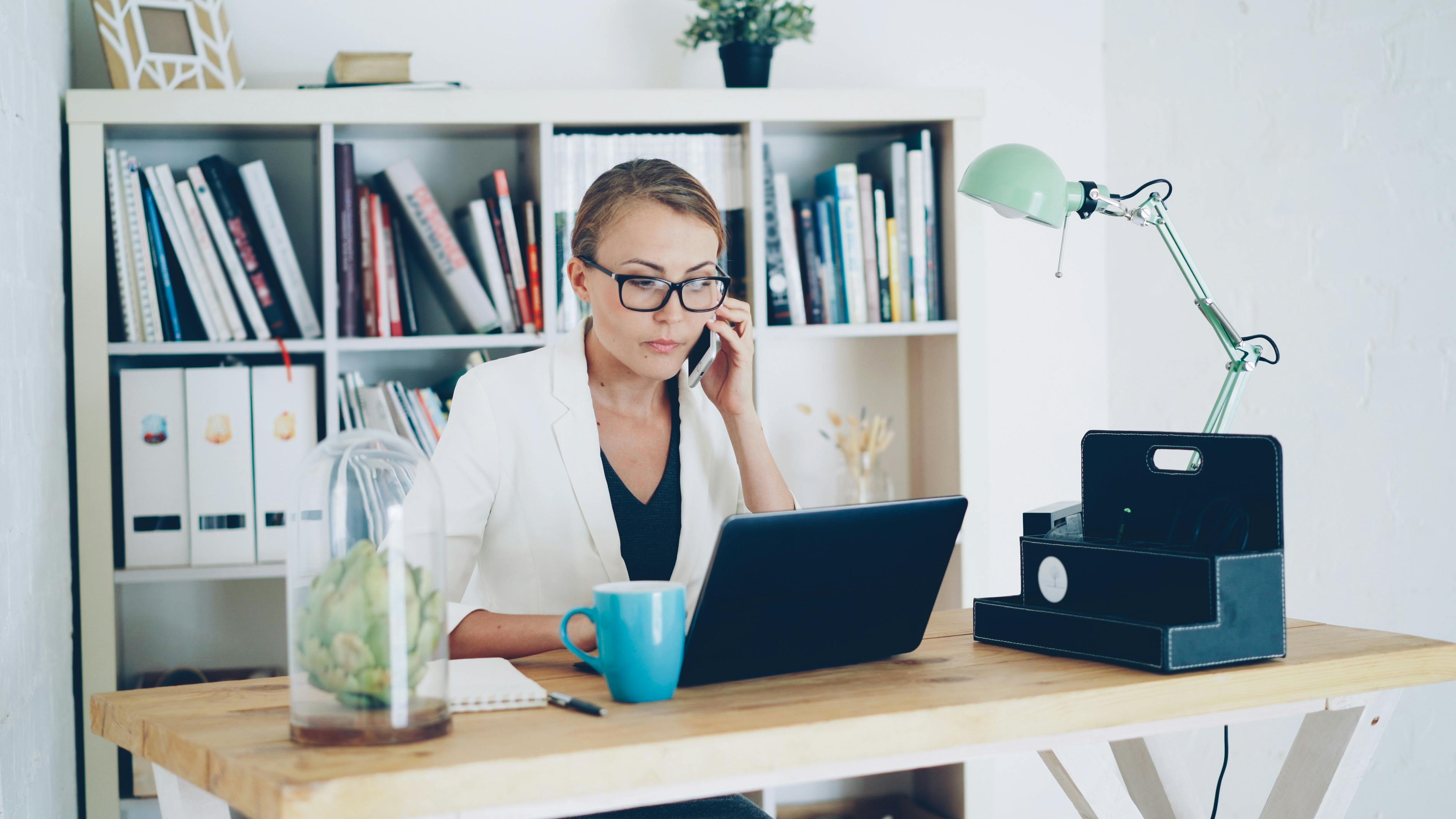 Businesswoman in office setting working on laptop while on a phone call, showcasing productivity.