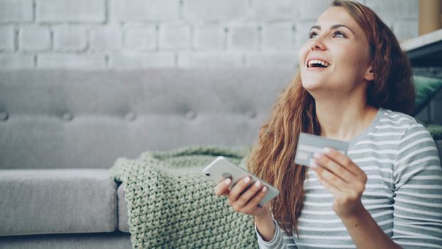 Joyful woman holding smartphone and credit card indoors, smiling at online shopping success.