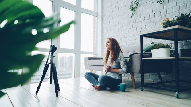 Young woman meditating in a bright room, facing a camera on a tripod, surrounded by plants.