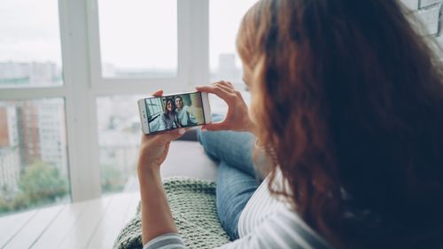 Free A woman captures a selfie with friends on her smartphone while relaxing by a large window indoors. Stock Photo