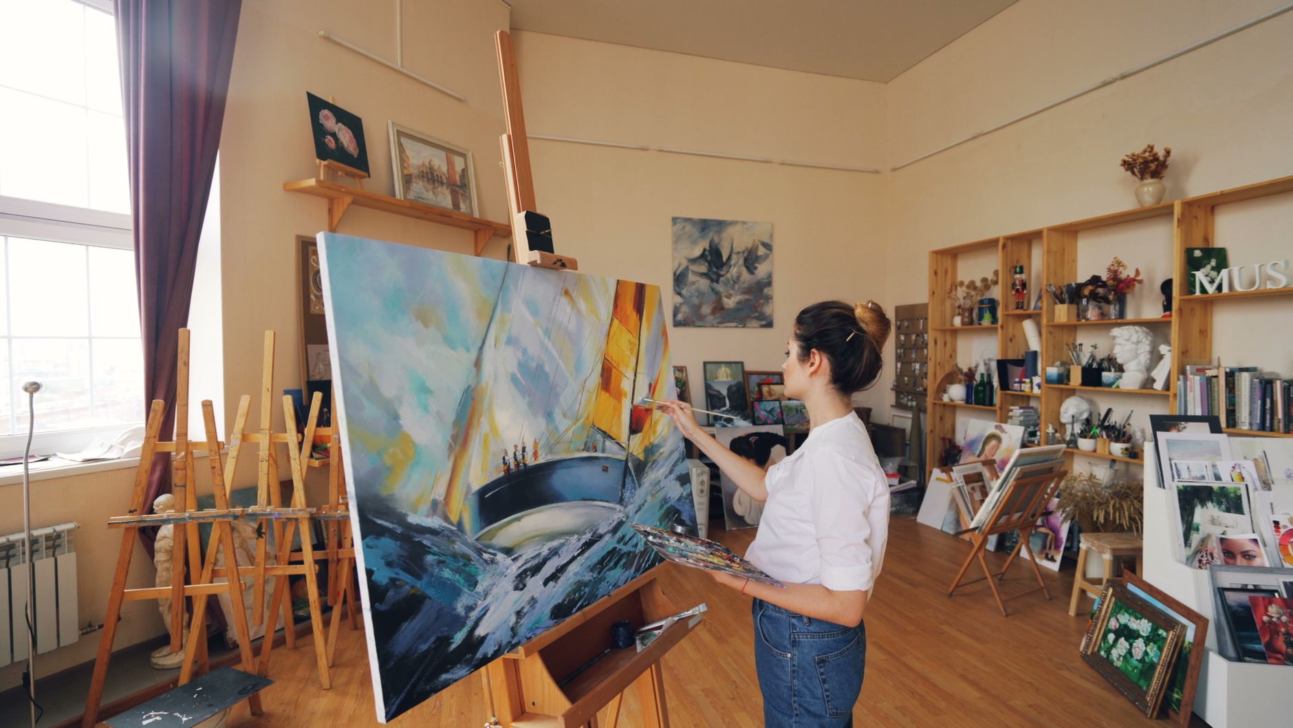 A female artist painting a colorful canvas in a sunlit art studio filled with creative supplies.