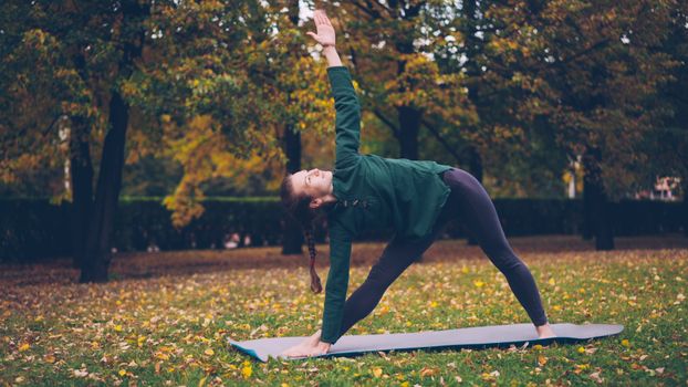 A woman executes a yoga pose on a mat in a scenic autumn park, surrounded by trees.