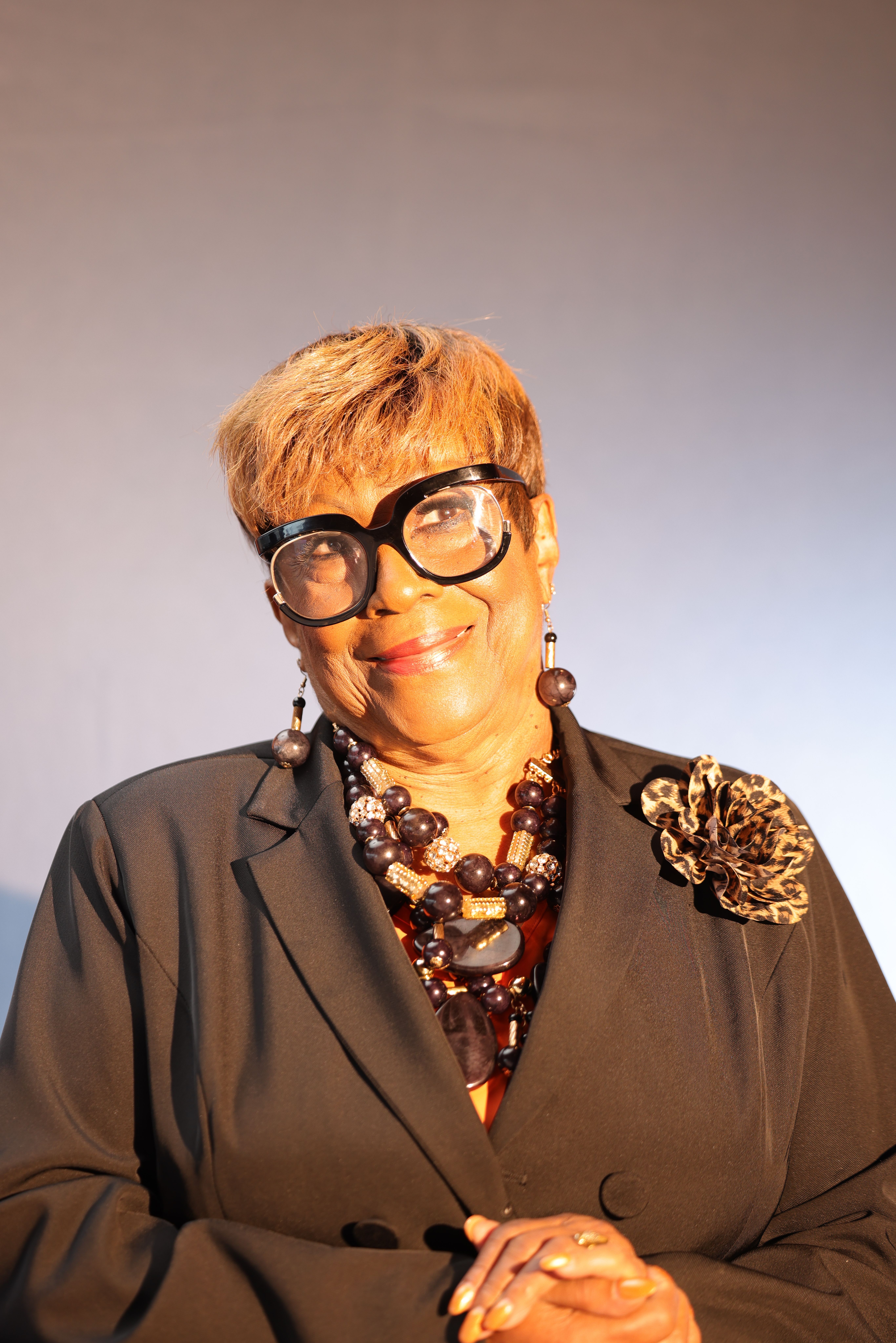 Elegant senior woman in stylish attire, featuring bold glasses and a statement necklace, looking confident.