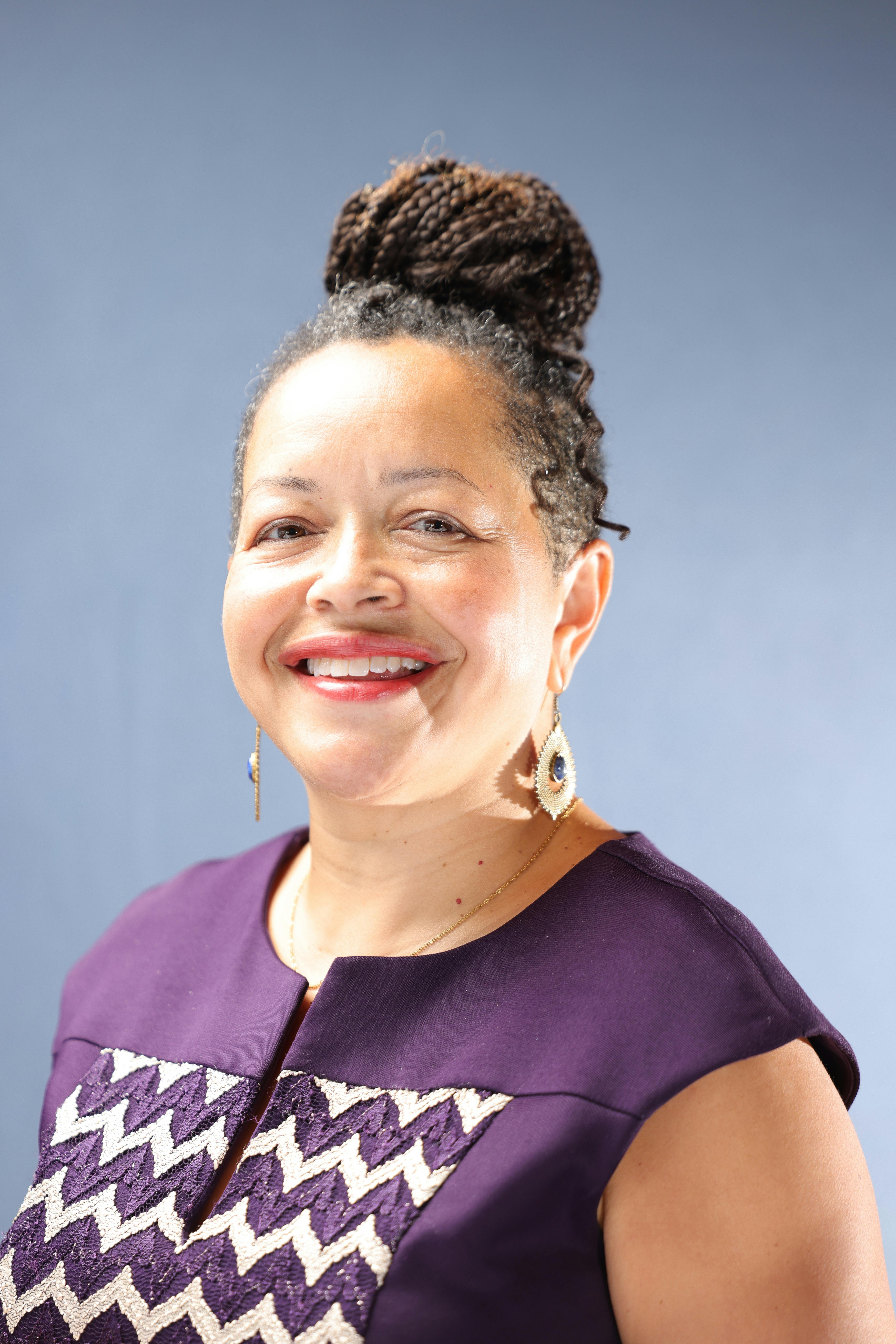 Portrait of a smiling woman with braided hair wearing a purple dress indoors.