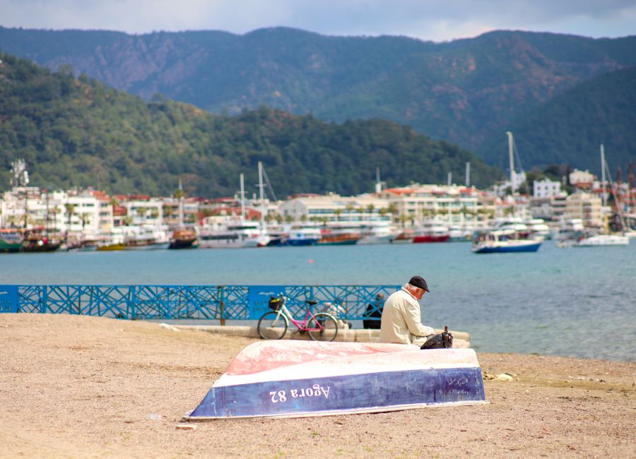 A serene coastal scene with a person seated by an overturned boat, overlooking the harbor and distant mountains.