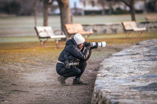 Free Photographer kneeling outdoors using a telephoto lens on a camera. Capturing moments in a serene park setting. Stock Photo
