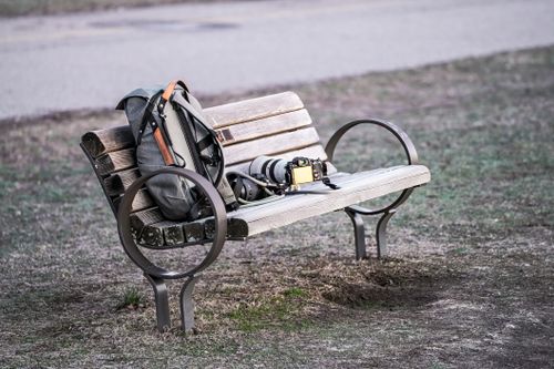 Free Camera and backpack placed on a park bench outdoors, exemplifying a travel photographer's break. Stock Photo