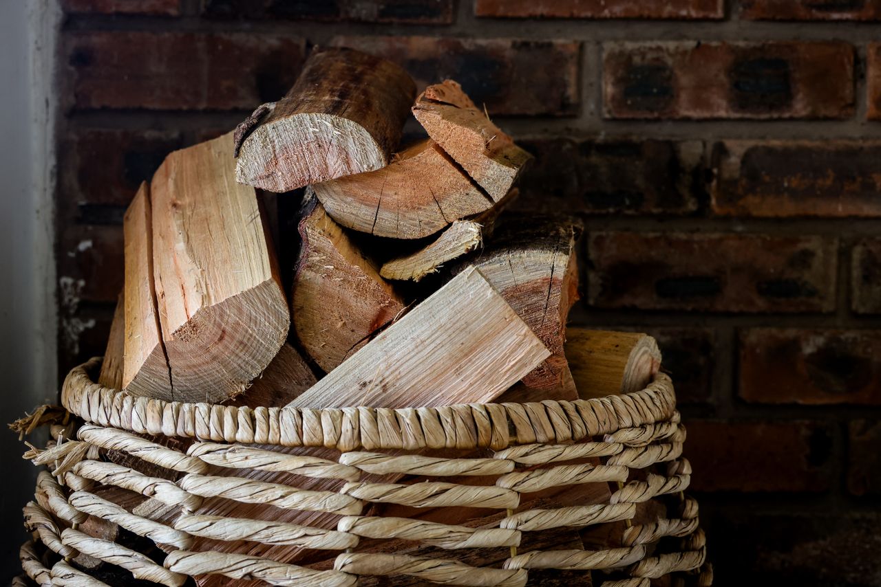 Woven basket filled with chopped firewood, including pale birch logs, against a brick wall.