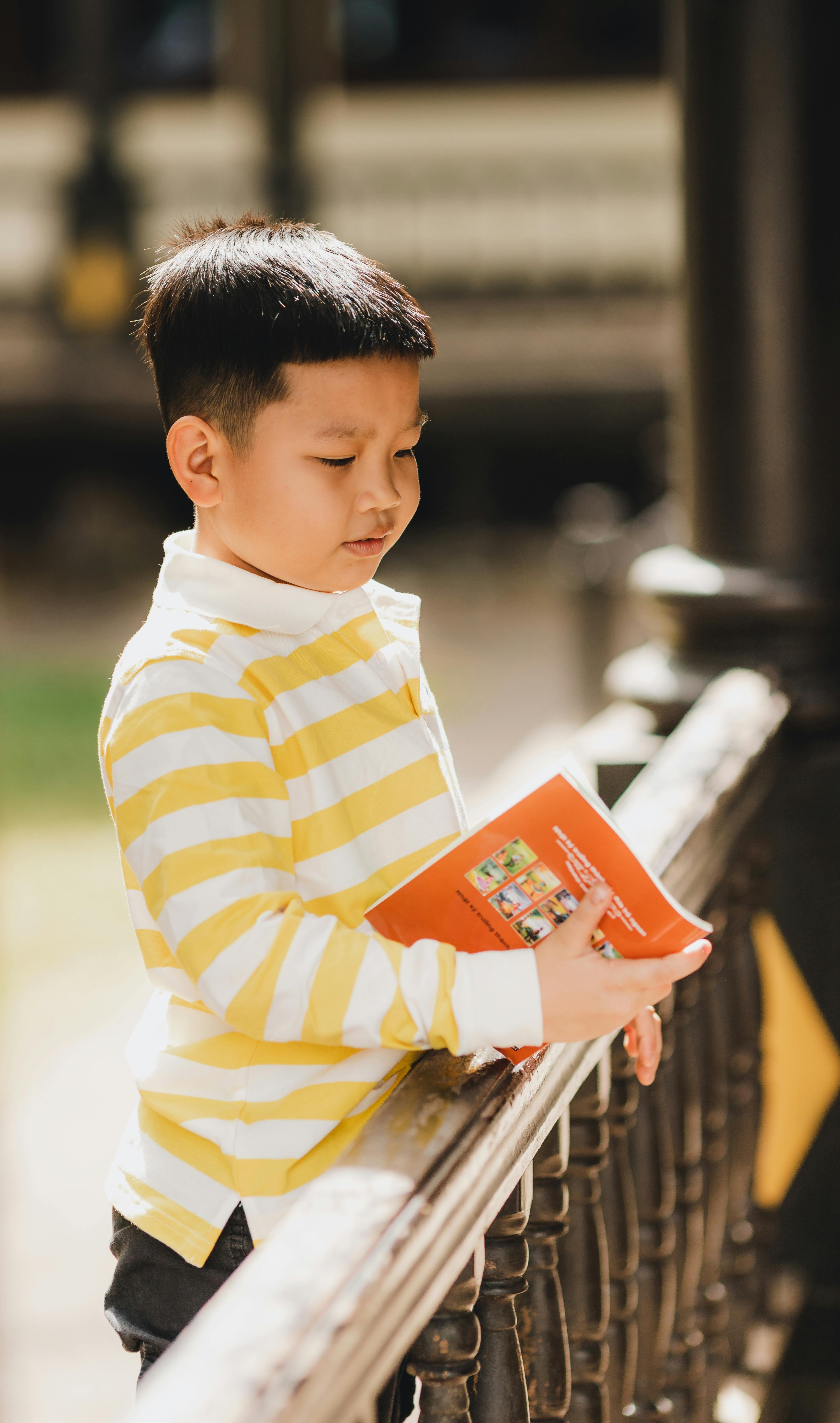 A young boy in a striped shirt reads a book while outdoors in Kon Tum, Vietnam, capturing a peaceful moment.
