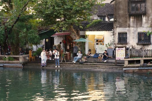 Serene riverside with people relaxing by traditional shops in a picturesque Chinese setting.