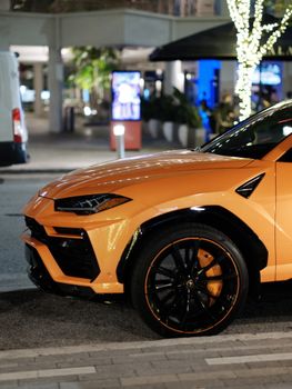 Vibrant orange luxury car parked on a city street at night with decorative lighting.