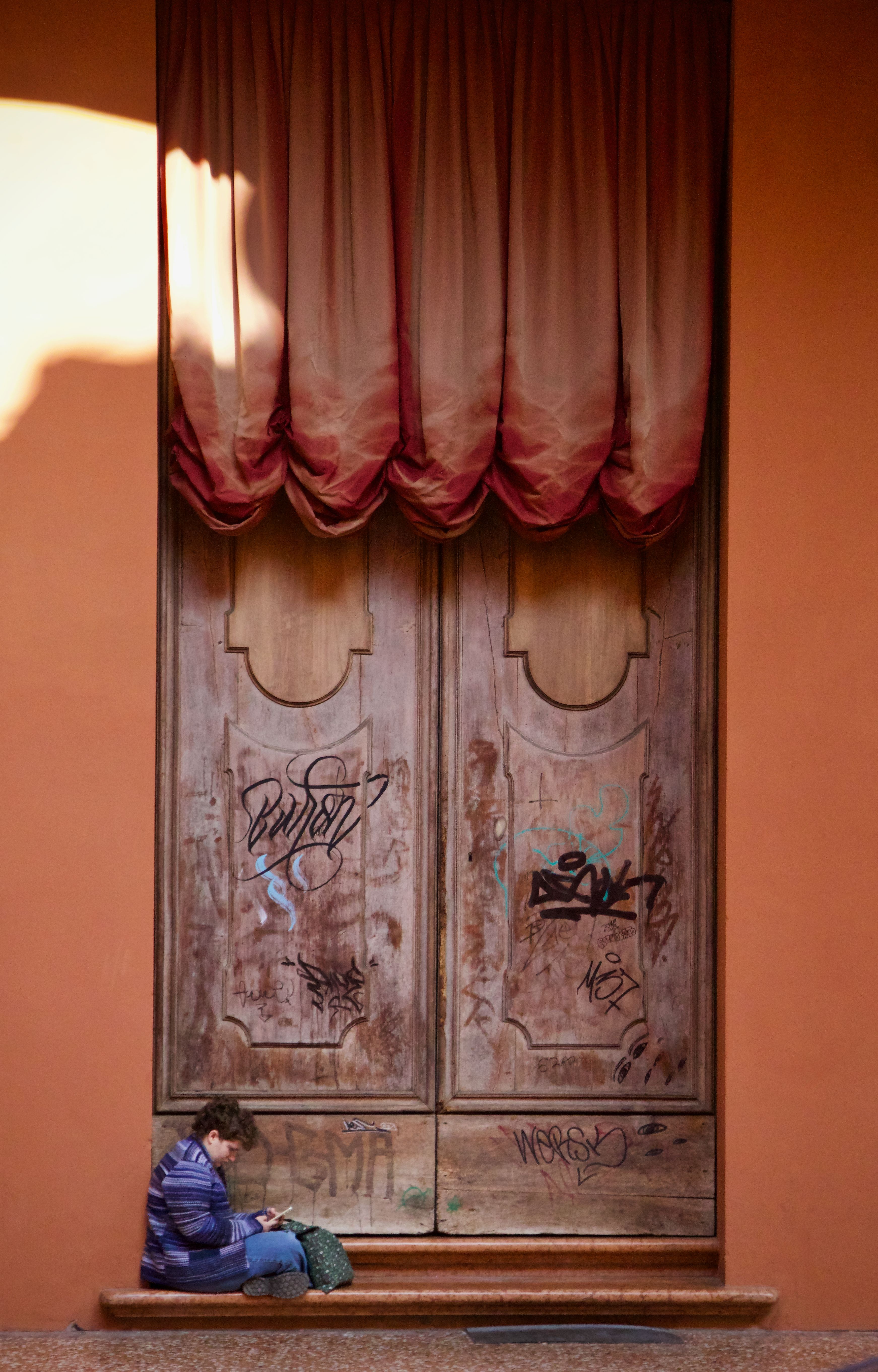 A child in a purple jacket sits by ornate wooden doors with graffiti in warm sunlight.