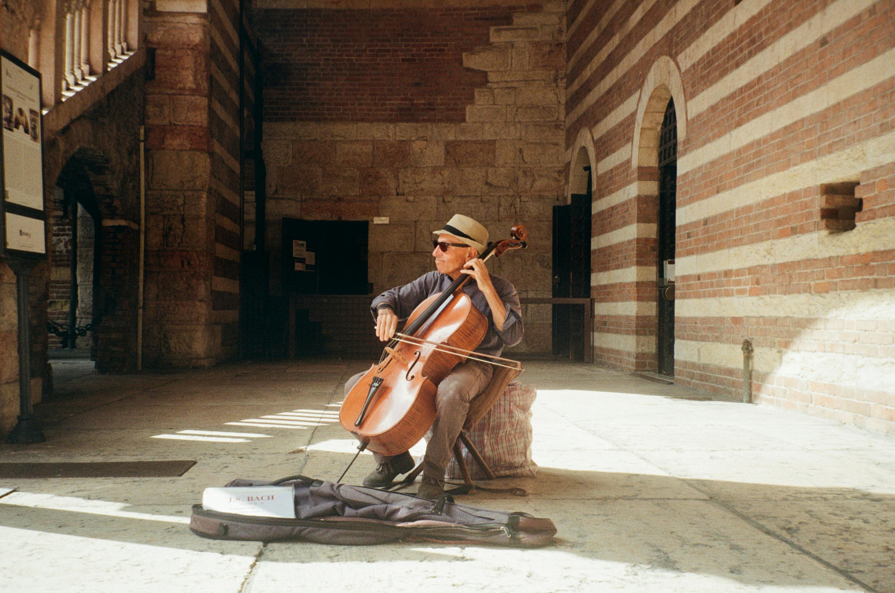 A street musician plays the cello in a sunlit historic archway, wearing a hat and sunglasses.