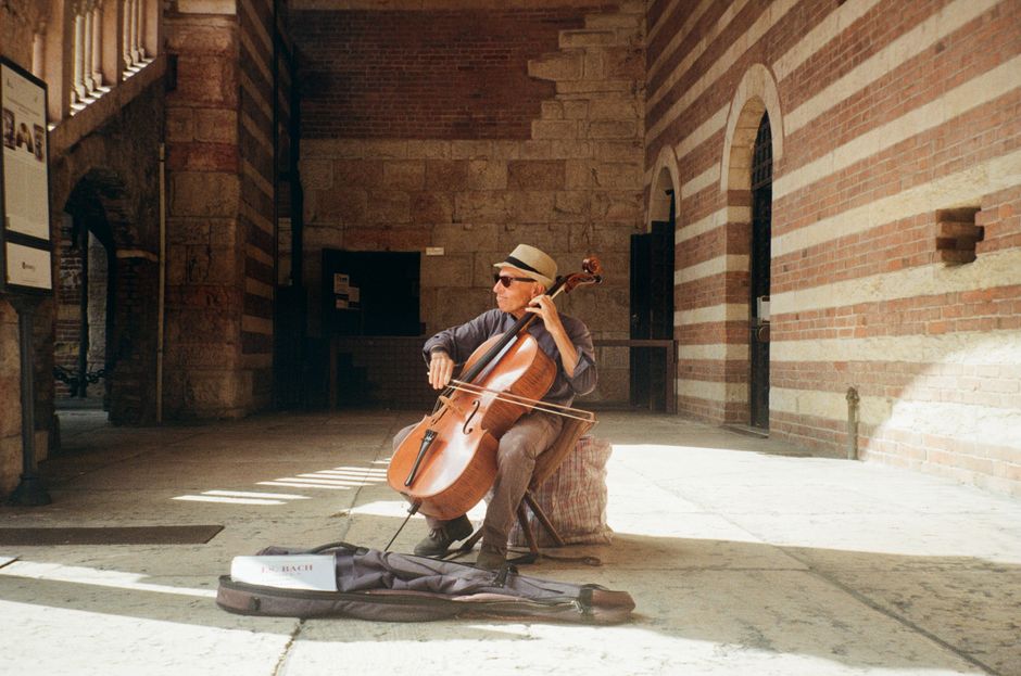 A street musician plays the cello in a sunlit historic archway, wearing a hat and sunglasses.