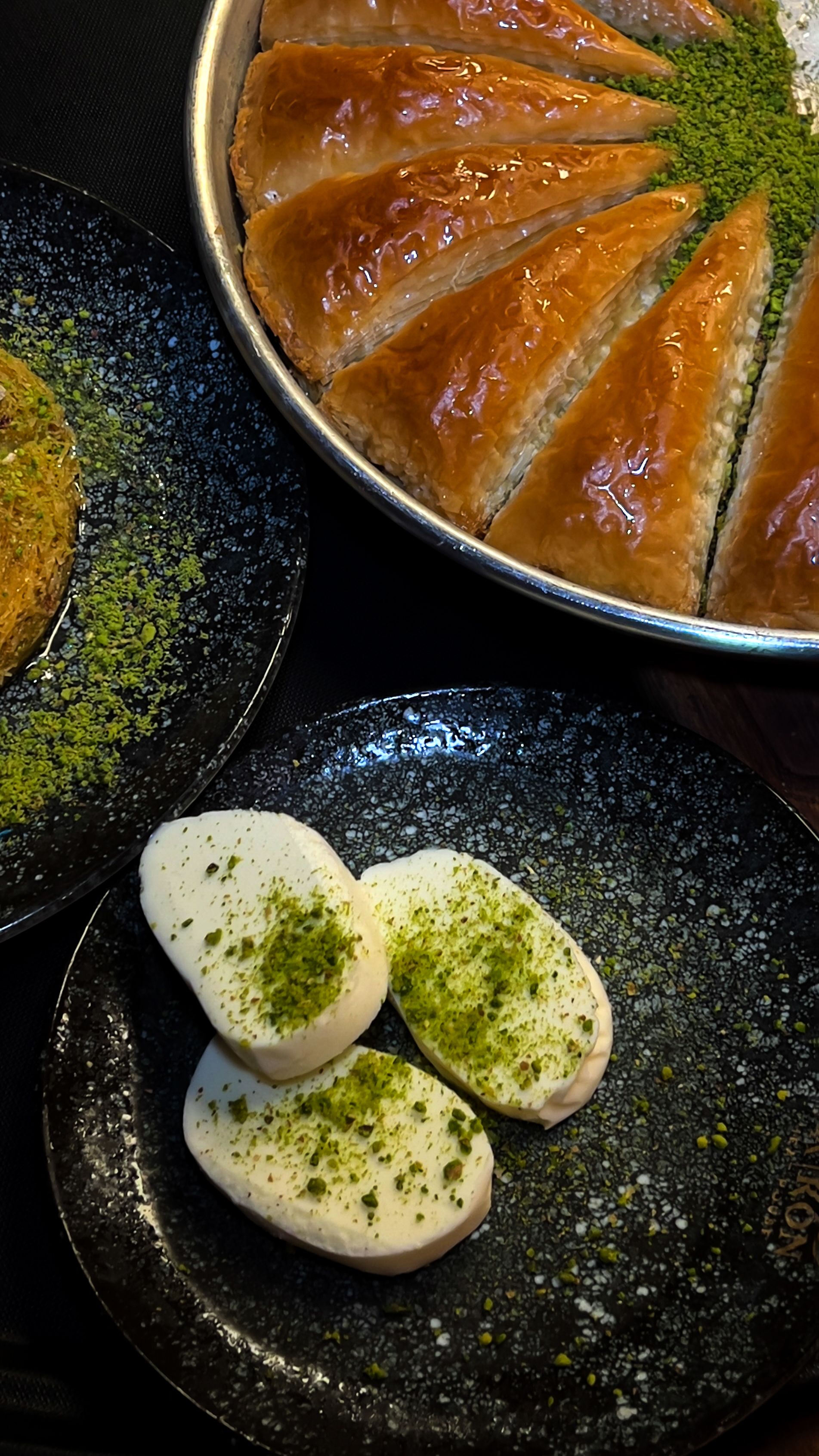 Close-up of Middle Eastern pastries and desserts topped with pistachios on a black plate.