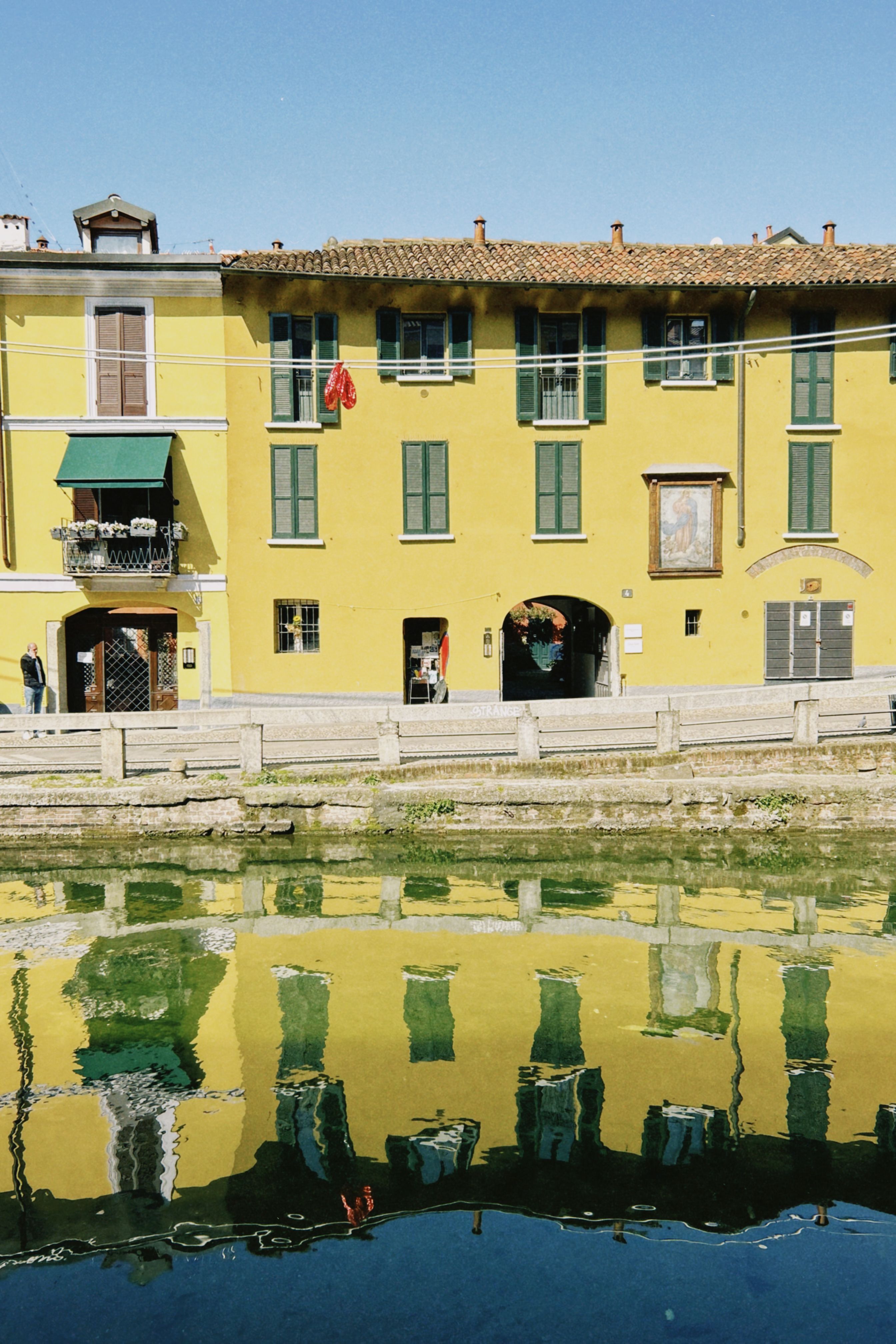 Yellow building with green windows reflects in a canal, creating a colorful scene on a sunny day.
