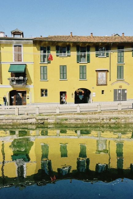 Yellow building with green windows reflects in a canal, creating a colorful scene on a sunny day.