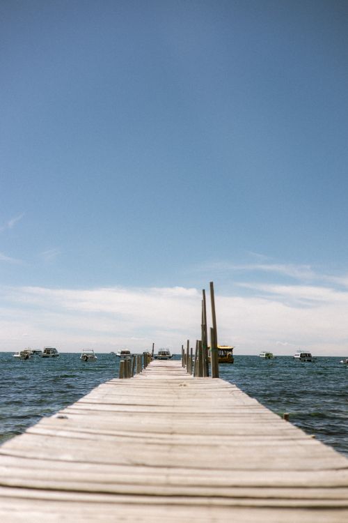 Free A serene view of a wooden pier extending into Lake Titicaca with boats in the distance under a clear blue sky. Stock Photo