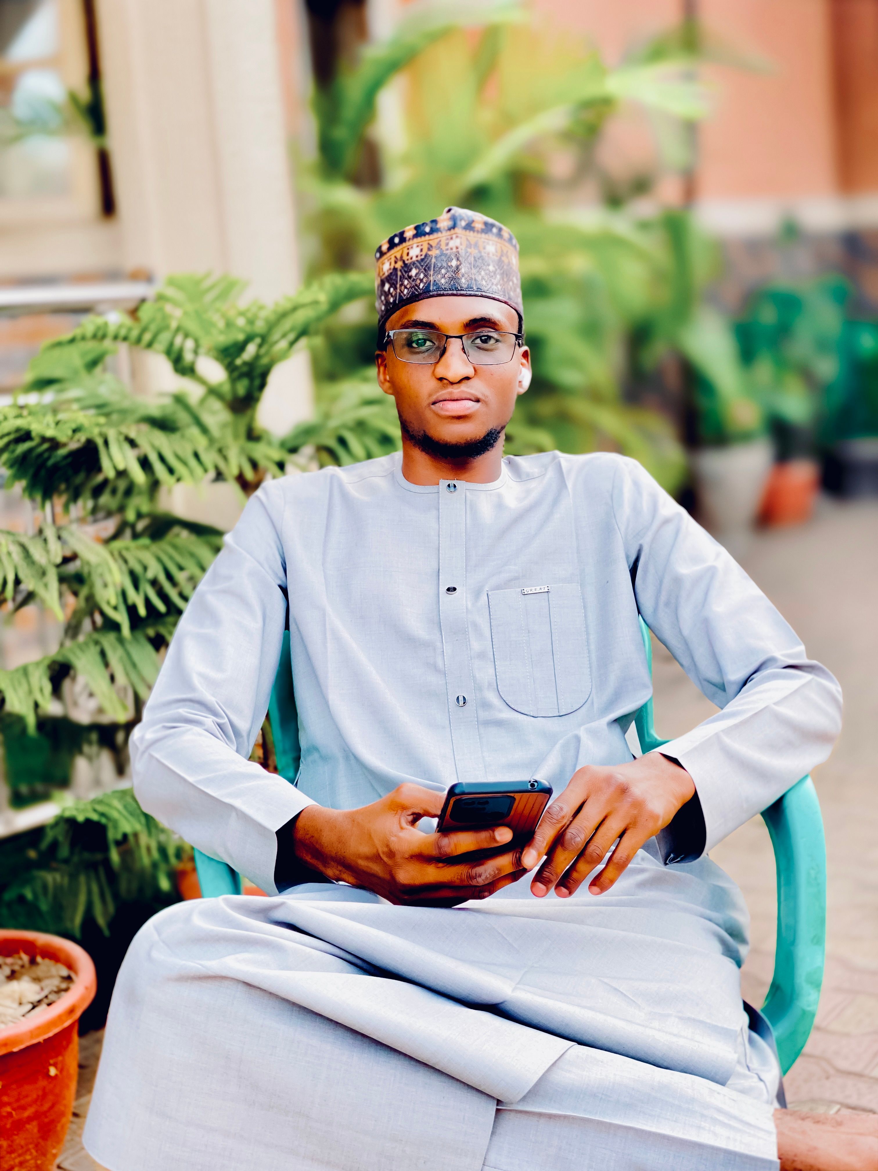 Young man in traditional attire seated outdoors with smartphone, surrounded by greenery.